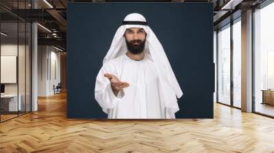 Half-length portrait of arabian saudi businessman on dark blue studio background. Young male model smiling, showing a gesture of inviting. Concept of business, finance, facial expression, human Wall mural