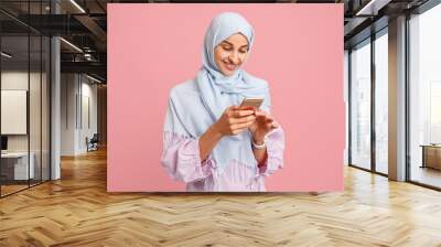 Happy arab woman in hijab with mobile phone. Portrait of smiling girl, posing at pink studio background. Young emotional woman. The human emotions, facial expression concept. Front view. Wall mural