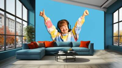 Happy smiling teen girl with down syndrome listening to music in headphones, expressing joy against blue background. Concept of acceptance, care, inclusion, health, diversity, emotions, equality Wall mural