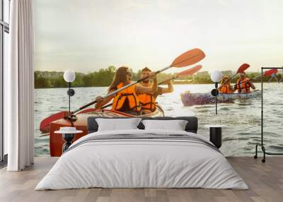 Happy young caucasian group of friends kayaking on river with sunset in the backgrounds. Having fun in leisure activity. Happy male and female model laughting on the kayak. Sport, relations concept. Wall mural