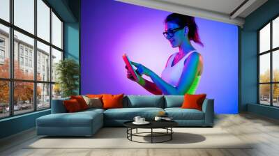 Holding tablet, crazy excited. Caucasian young woman's portrait on black studio background in neon light. Beautiful male model in white shirt. Wall mural