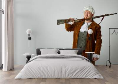 Hunter. Studio shot of young man in image of medieval person in vintage brown hunting suit and white wig with old hunting rifle isolated over grey background. Wall mural