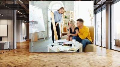 Interior designer working with young couple. Lovely family and professional designer or architector discussing conept of future interior, working with colour palette, room drawings in modern office. Wall mural