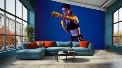 Kicking. Fit caucasian woman in sportswear boxing on blue studio background in neon light. Novice female caucasian boxer working out and training. Sport, healthy lifestyle, movement concept. Wall mural