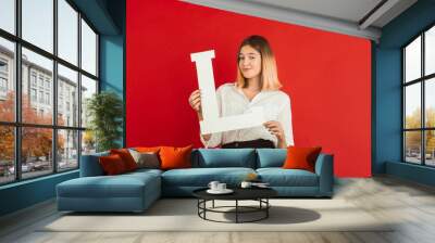 L - is Love. Valentine's day celebration, happy, cute caucasian girl holding letter on red studio background. Concept of human emotions, facial expression, love, relations, romantic holidays. Wall mural