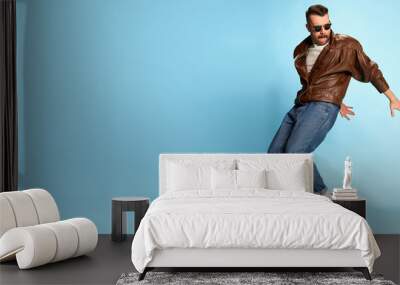 Portrait of brutal, handsome man in jeans, leather jacket and sunglasses posing, dancing over blue studio background. Banner, flyer. Concept of emotions, lifestyle Wall mural