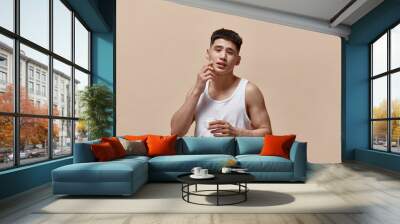 Portrait of handsome young man with clean, spotless face applying face moisturizing cream against light brown studio background. Concept of male beauty, skincare, cosmetology, men's health Wall mural