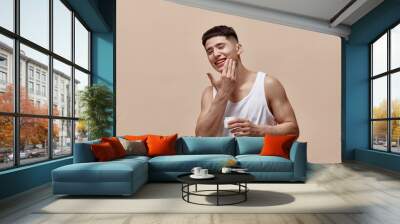 Portrait of smiling, handsome, young guy with clean, spotless face applying face moisturizing cream against light brown studio background. Concept of male beauty, skincare, cosmetology, men's health Wall mural