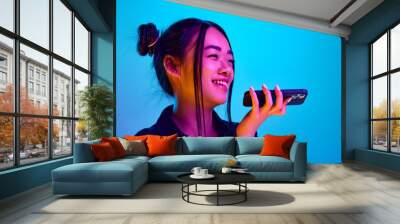 Portrait of young, happy, korean girl smiling and recording voice message on phone against blue studio background in neon light. Concept of emotions, facial expression, youth, lifestyle, inspiration Wall mural