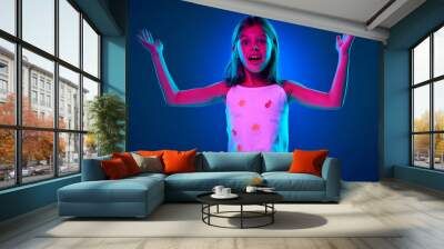 Portrait of young emotional girl in yellow polo shirt expressing shock, surprise and excitement against blue studio background in neon light. Concept of childhood, kids emotions, lifestyle Wall mural