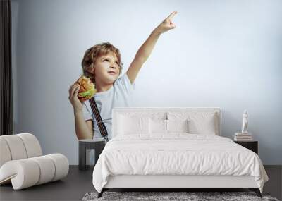 Pretty young curly boy in casual clothes on white studio background. Eating burger. Caucasian male preschooler with bright facial emotions. Childhood, expression, fun, fast food. Dreamful looks up. Wall mural