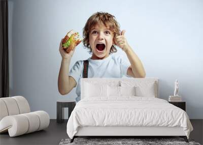 Pretty young curly boy in casual clothes on white studio background. Eating burger. Caucasian male preschooler with bright facial emotions. Childhood, expression, fun, fast food. Showing thumb up. Wall mural