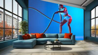 Ropes. Young sportive girl training with sports equipment isolated on gradient blue-pink studio background in neon light. Sport, action, fitness, youth concept. Wall mural