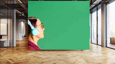 Senior man and woman standing back to back, listening to music in headphones against green studio background. Concept of beauty and fashion, relationship, modern style, age. Banner Wall mural