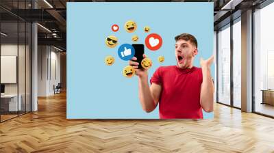 Social media interactions on mobile phone. Internet digital marketing, Chating, commenting, liking. Smiles and icons above smartphone screen, that holding by young man on blue studio background. Wall mural