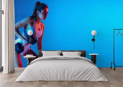 Strength training. Young sportive girl training with sports equipment isolated on gradient blue-pink studio background in neon light. Sport, action, fitness, youth concept. Wall mural