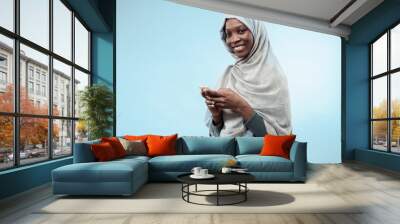 The beautiful young black african muslim girl wearing gray hijab at blue studio. She standing with mobile phone with a happy smile on her face. The human emotions, facial expression concept. Trendy Wall mural