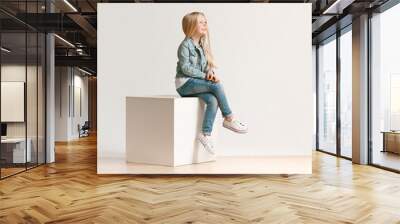 The portrait of cute little kid girl in stylish jeans clothes looking at camera and smiling, sitting against white studio wall. Kids fashion concept Wall mural