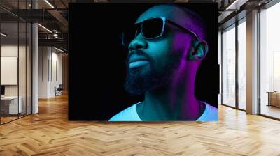 The retro wave or synth wave portrait of a young happy smiling african man in sunglasses at studio. High Fashion male model in colorful bright neon lights posing on black background. Art design Wall mural