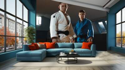 Two young judo caucasian fighters in white and blue kimono with black belts posing confident in the gym, strong and healthy. Practicing martial arts fighting skills. Overcoming, reaching target. Wall mural