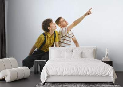 Two young men sitting on white cube, one pointing upwards with excitement, other looking in awe, capturing moment of discovery. News, sales. Concept of youthful curiosity, announcement Wall mural