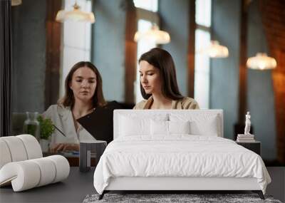 Two young women in formal wear sitting at table and checking business papers. Project preparation. Business meeting in office. Concept of business, teamwork, career development, brainstorming Wall mural