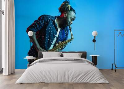 Young african-american jazz musician playing the saxophone on blue studio background in trendy neon light. Concept of music, hobby. Joyful attractive guy improvising. Retro colorful portrait of artist Wall mural