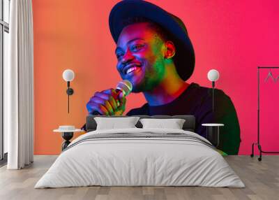 Young african-american musician singing on gradient orange-purple studio background in neon light. Concept of music, hobby, festival. Joyful party host, stand upper. Colorful portrait of artist. Wall mural