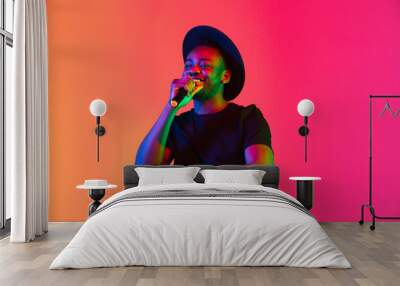 Young african-american musician singing on gradient orange-purple studio background in neon light. Concept of music, hobby, festival. Joyful party host, stand upper. Colorful portrait of artist. Wall mural