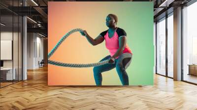 Young african-american plus size female model's training on gradient background in neon light. Doing workout exercises with the ropes. Concept of sport, healthy lifestyle, body positive, equality. Wall mural