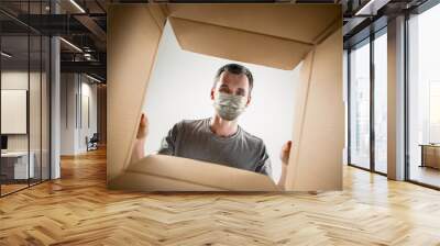 Young man opening the huge postal package wearing protective face mask. Male model on top of cardboard box. Food and goods contactless delivery during coronavirus quarantine for isolated people. Wall mural