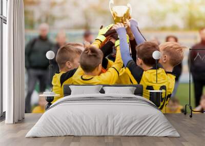 Happy kids in yellow shirts in elementary school sports team celebrating soccer succes in tournament final game. Sports team members raising golden cup trophy Wall mural