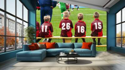 School soccer team witch coach on tournament match sitting on wooden bench. Children in red soccer shirts ready to play football game Wall mural