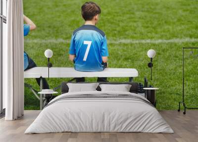 Schoolboy sitting on soccer bench. Young boy sitting on the substitute bench. Football sports competition game for children. The little boy in a blue jersey with the number seven Wall mural