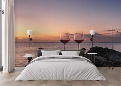 Two glasses of red wine  on the  rock beach near water. Romantic date on a beautiful sunset ocean background.  Romantic glasses of red wine against a colorful sunset. Wall mural