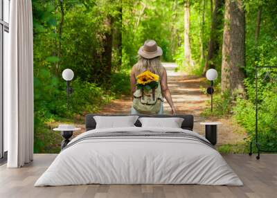 A blonde caucasian woman walks down a trail in the forest with sunflowers sticking out of her backpack on a sunny Summer's Day  Wall mural