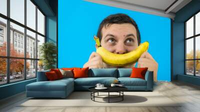 Giovane ragazzo si diverte creando un sorriso con una banana su sfondo blu Wall mural