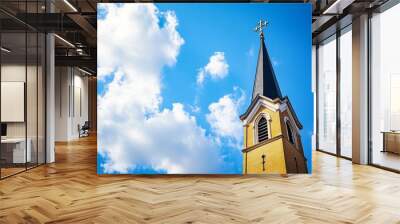 A tall church steeple with a cross against a bright blue sky and fluffy clouds. Wall mural