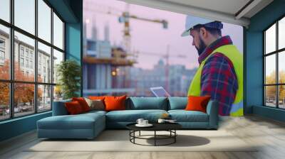 engineer in a white helmet and yellow vest standing on a construction site roof with cranes, using a tablet Wall mural