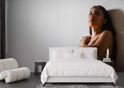 brunette latina woman covering her breasts with her hands folded while leaning against the wall with a sad look looking up. Wall mural