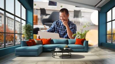 Smiling Asian designer working on a laptop in an office Wall mural