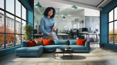 Smiling businesswoman sitting on an office table holding a tablet Wall mural