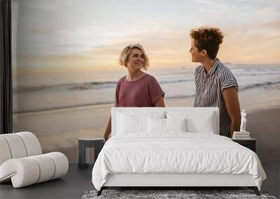 Smiling lesbian couple walking along a beach at sunset Wall mural