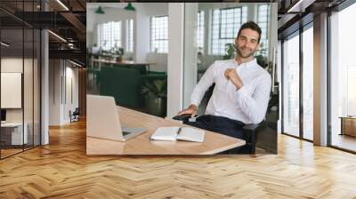 Smiling young businessman sitting at his desk in an office Wall mural