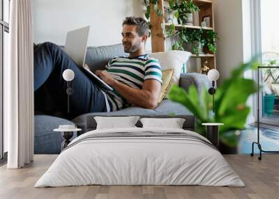 Smiling young man using a laptop on his sofa Wall mural