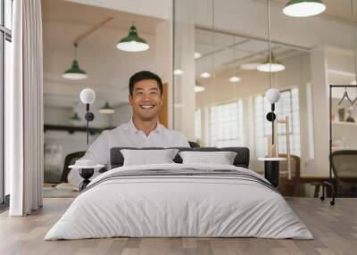 Successful Asian businessman smiling while sitting at his office desk Wall mural