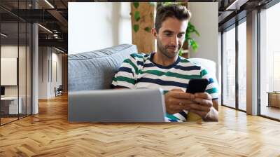 Young man using a cellphone and laptop on his sofa Wall mural