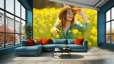 A woman in a green floral dress enjoys a sunny day in a vibrant yellow flower field near a forest during springtime. Nature, fashion, summer lifestyle. Wall mural