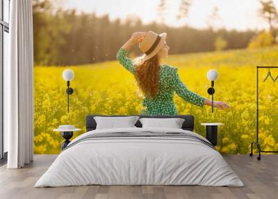 Beautiful woman in bright dress and elegant hat walks and has fun in rapeseed field. Smiling female tourist walking through flowering field, touching yellow flowers. Nature, rest. Summer landscape. Wall mural