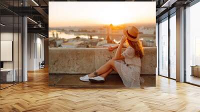 Beautiful woman in hat enjoys stunning view of the city at dawn. Back view. Tourist is looking at landscape of city from height. Travel in Europe. Wall mural